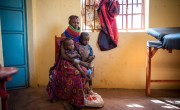Woman wearing colourful necklaces sits in doctor waiting room with her two infant sons on her lap