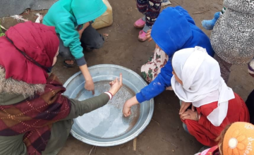 Concern staff, wearing a black facemask, demonstrating the importance of using soap when washing hands and explaining the importance of the critical times to wash hands. To do so chilli is used to demonstrate if no soap is used the chilli flakes remain on the hands, whereas when soap is used they are removed from the hands and remain in the bowl. Syria, 2020. Photo: Concern Worldwide.