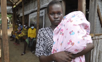 Woman stands outside health centre with baby swathed in a pink blanket in her arms. Four people sit behind her wearing face coverings.