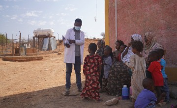 Doctor speaking to family with group of children, standing in shade next to building
