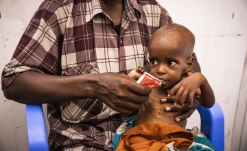 Thin infant eating from pack of therapeutic food held by man holding him