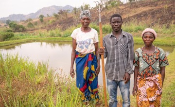 Man holding hoe in between two women, standing in front of pond