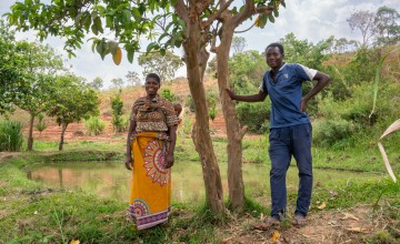 Man leaning against a tree and smiling, woman with baby on sling against her back smiling