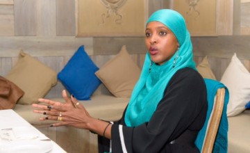 Activist Ifrah Ahmed speaking at table, wearing blue head scarf and black dress