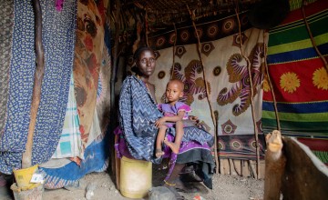 Lokaale Lorubun sitting in shelter surrounded by wall hangings, with child sitting on her lap