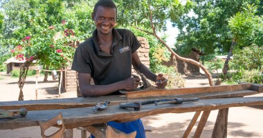 Man in dark t-shirt smiling and standing behind carpentry table 
