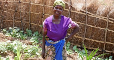 Elderly woman in purple t-shirt and green head wrap standing in garden with hand on hip 
