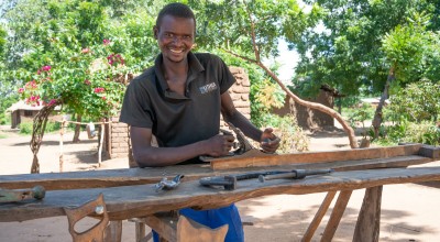 Man in dark t-shirt smiling and standing behind carpentry table 
