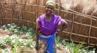 Elderly woman in purple t-shirt and green head wrap standing in garden with hand on hip 