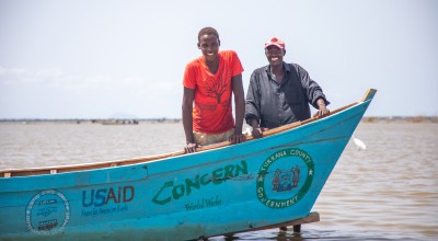 Two men stand in the sea behind a wooden fishing boat 