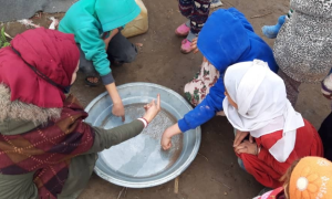 Concern staff, wearing a black facemask, demonstrating the importance of using soap when washing hands and explaining the importance of the critical times to wash hands. To do so chilli is used to demonstrate if no soap is used the chilli flakes remain on the hands, whereas when soap is used they are removed from the hands and remain in the bowl. Syria, 2020. Photo: Concern Worldwide.